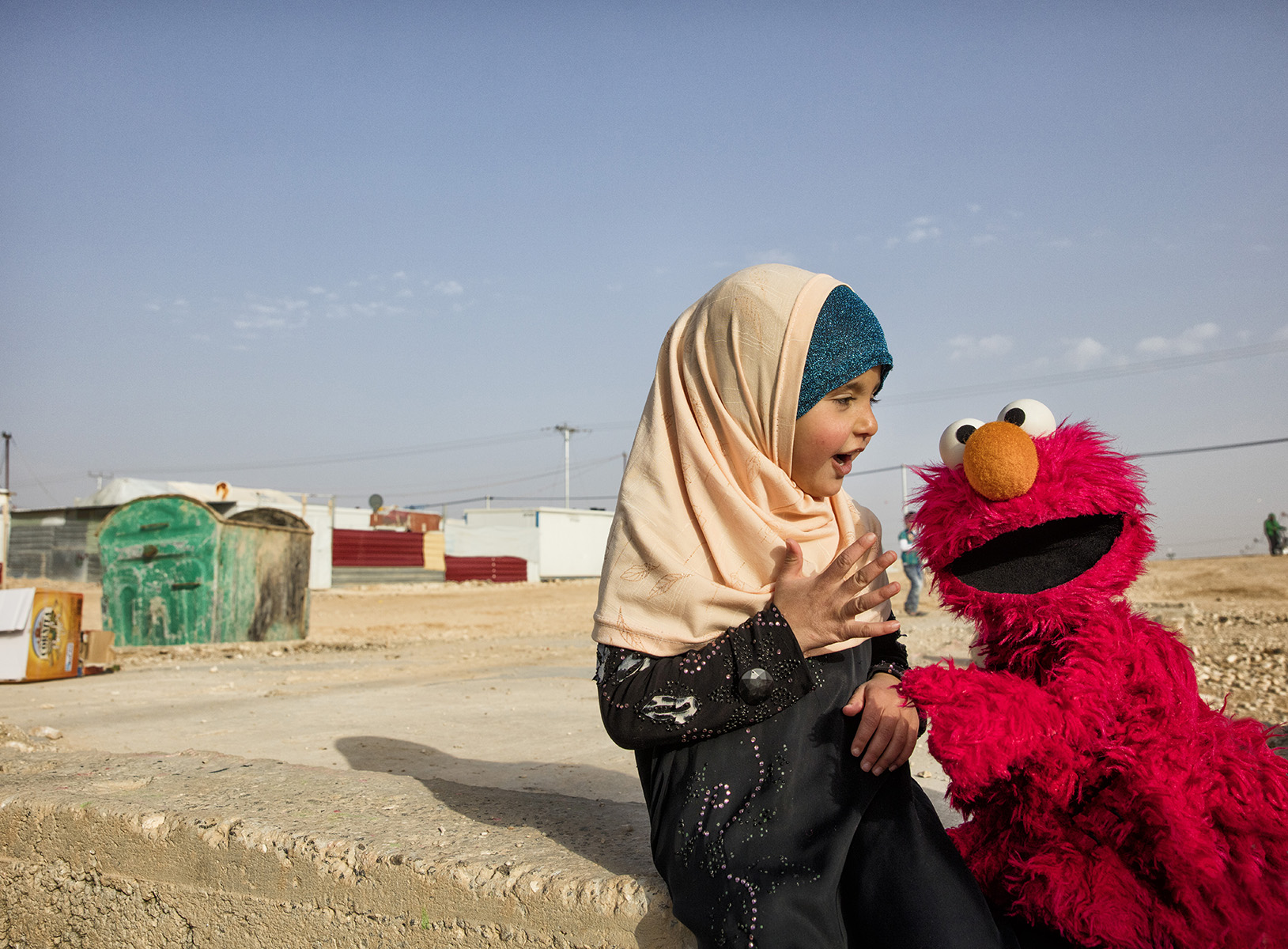 A young girl in a hijab sits outside, smiling and interacting with an Elmo puppet. The background shows a refugee camp with modest structures and clear skies.