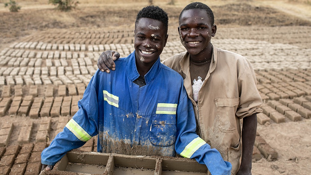 two men holding bricks in a dirt field.