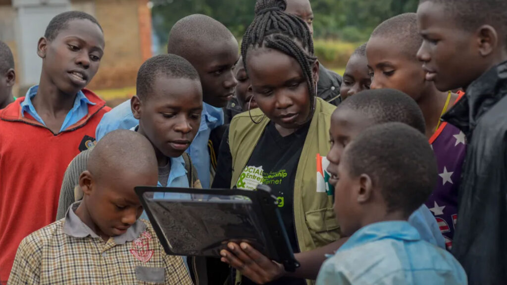 A woman holds a tablet, surrounded by a group of children and adolescents who are attentively looking at the screen. They appear to be outdoors in a rural setting.