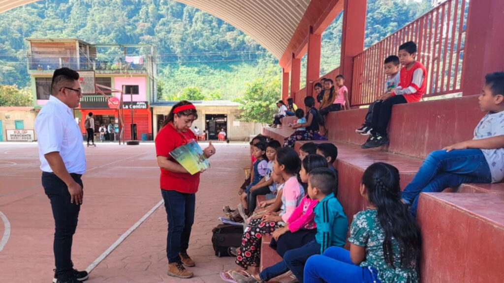 A group of children seated on an outdoor sports court listens attentively to a man reading a picture book. Another man stands nearby. Trees and buildings are visible in the background.