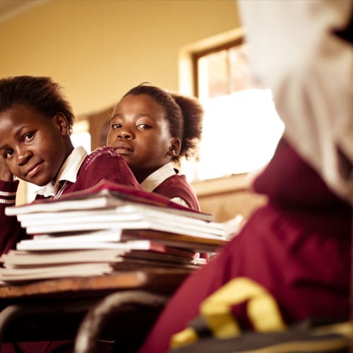 Two students in uniforms listening attentively in a classroom with a stack of papers in the foreground.