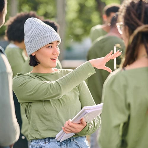 A person in a gray beanie gesturing while talking to another individual in a casual outdoor setting.