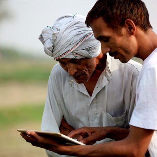 Two men, one older with a turban and one younger, looking at a tablet together outdoors.