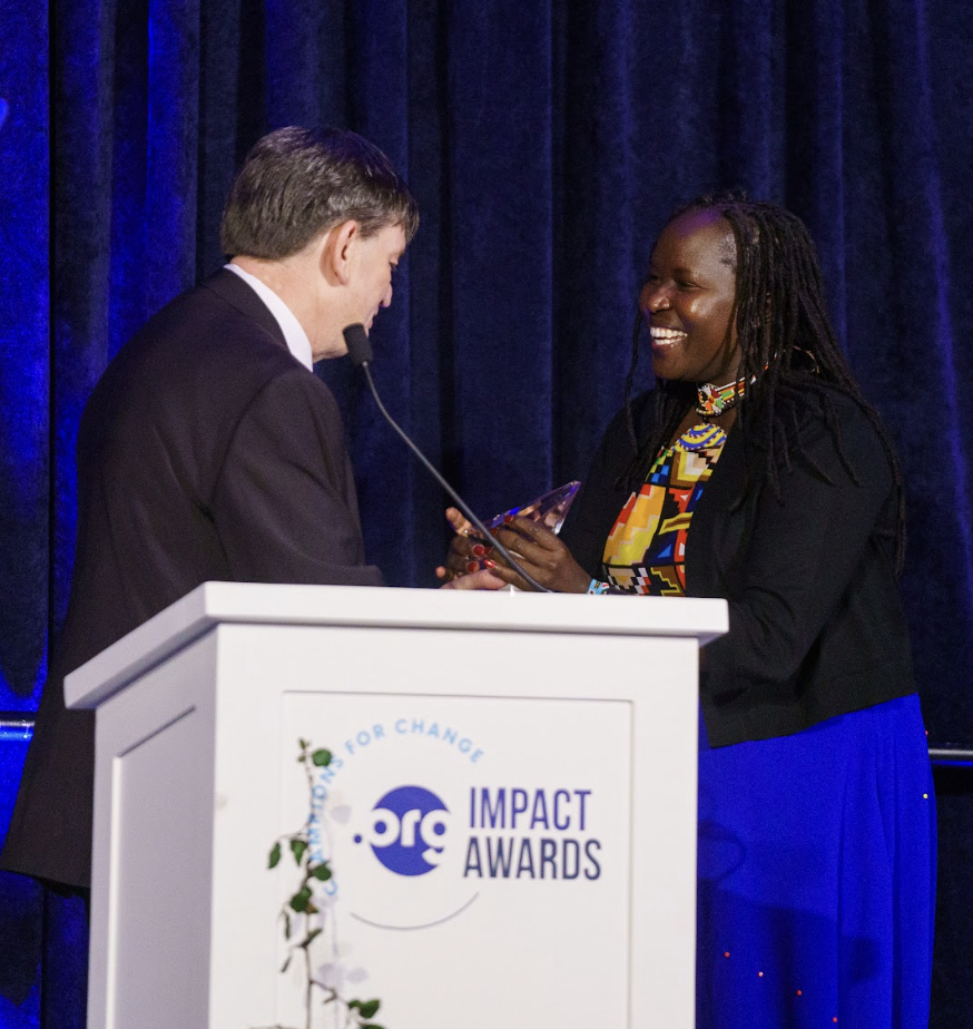 Two individuals, one presenting an award to the other, stand at a podium labeled ".ORG Impact Awards." The recipient is smiling while receiving the award.