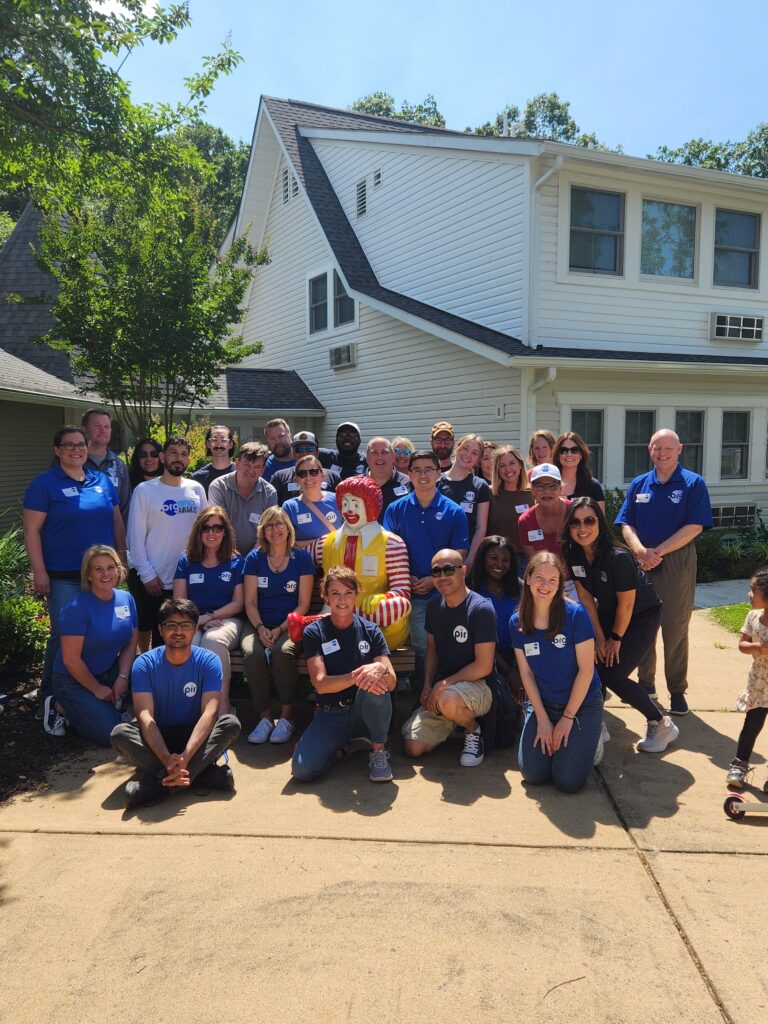 A group of people, including a person dressed as Ronald McDonald, pose for a photo outside a house. Most are wearing blue shirts and name tags.