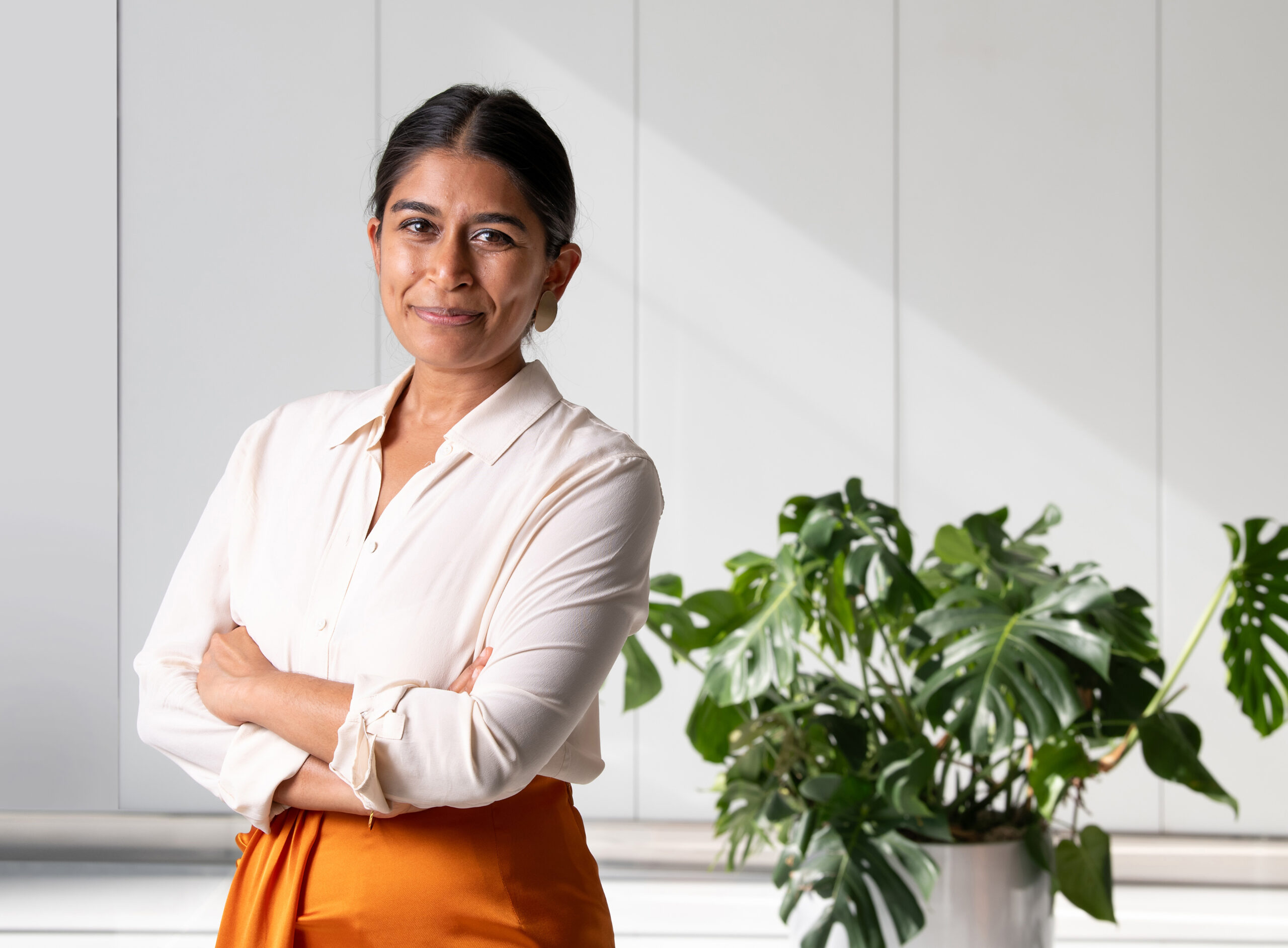 A woman in a white blouse stands with her arms crossed, smiling. She is in front of a white paneled wall with a potted plant beside her.