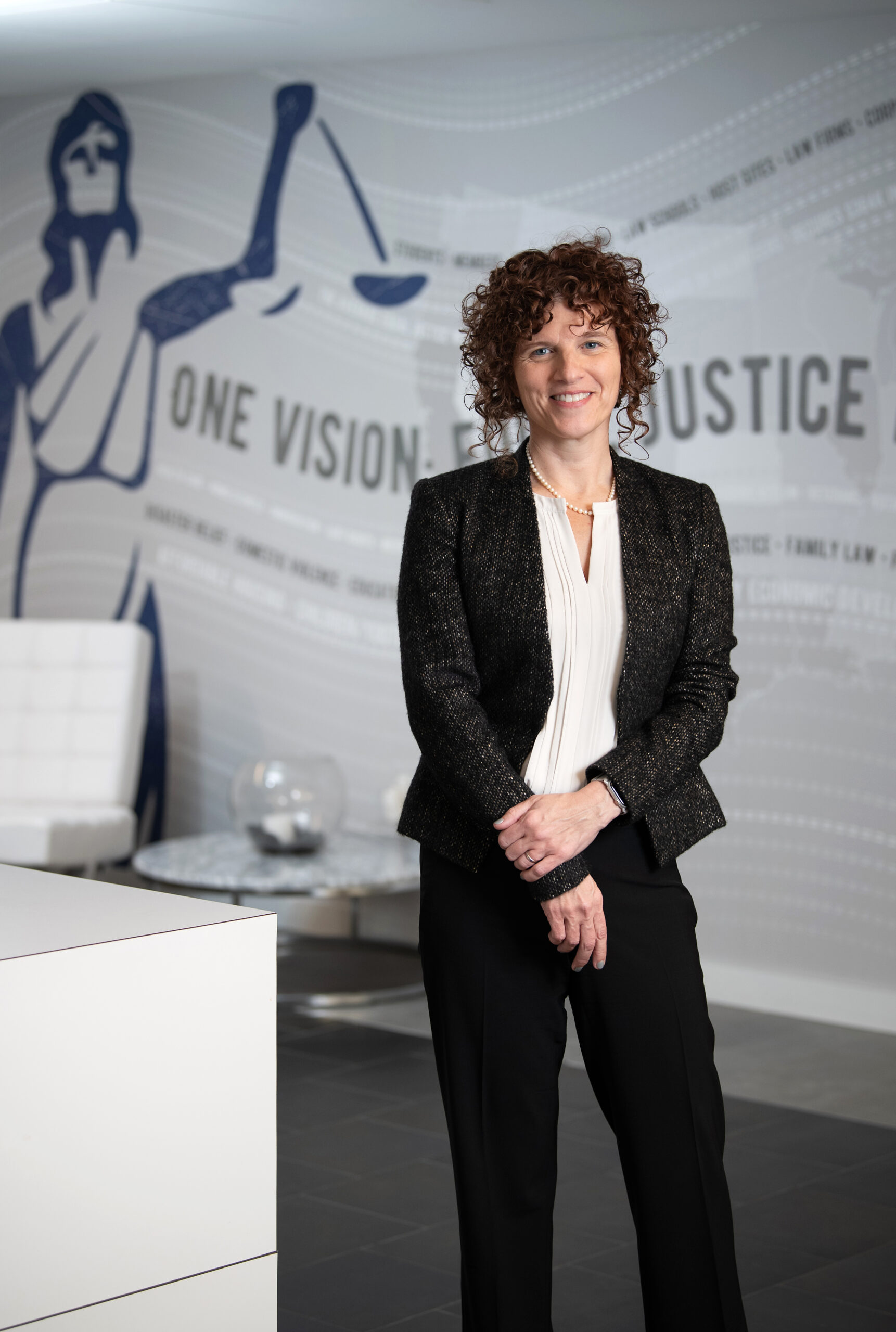 A woman with curly hair stands in an office, wearing a dark blazer and light blouse. Behind her is a wall mural featuring the scales of justice and the words "One Vision: Justice.