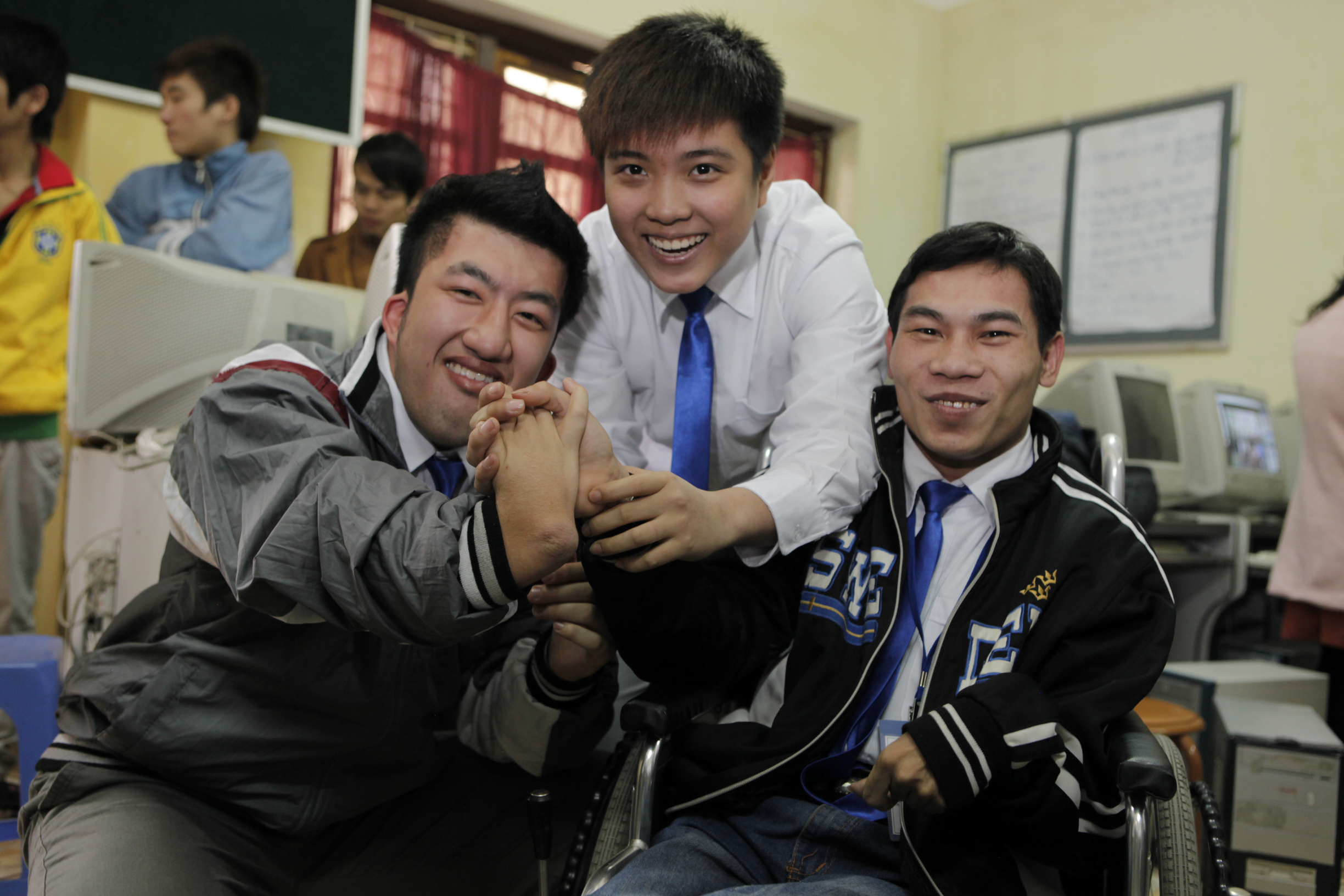 Three smiling men in a classroom, two sitting and one standing behind them, all wearing blue ties. Two of the men are clasping hands, and computers are visible in the background.