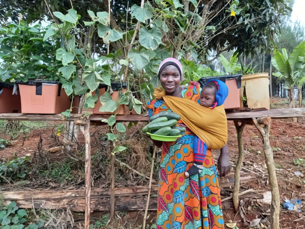 A woman stands outdoors holding a plate of cucumbers in one hand and carrying a child in a colorful wrap on her back, with vines and vegetable planters in the background.