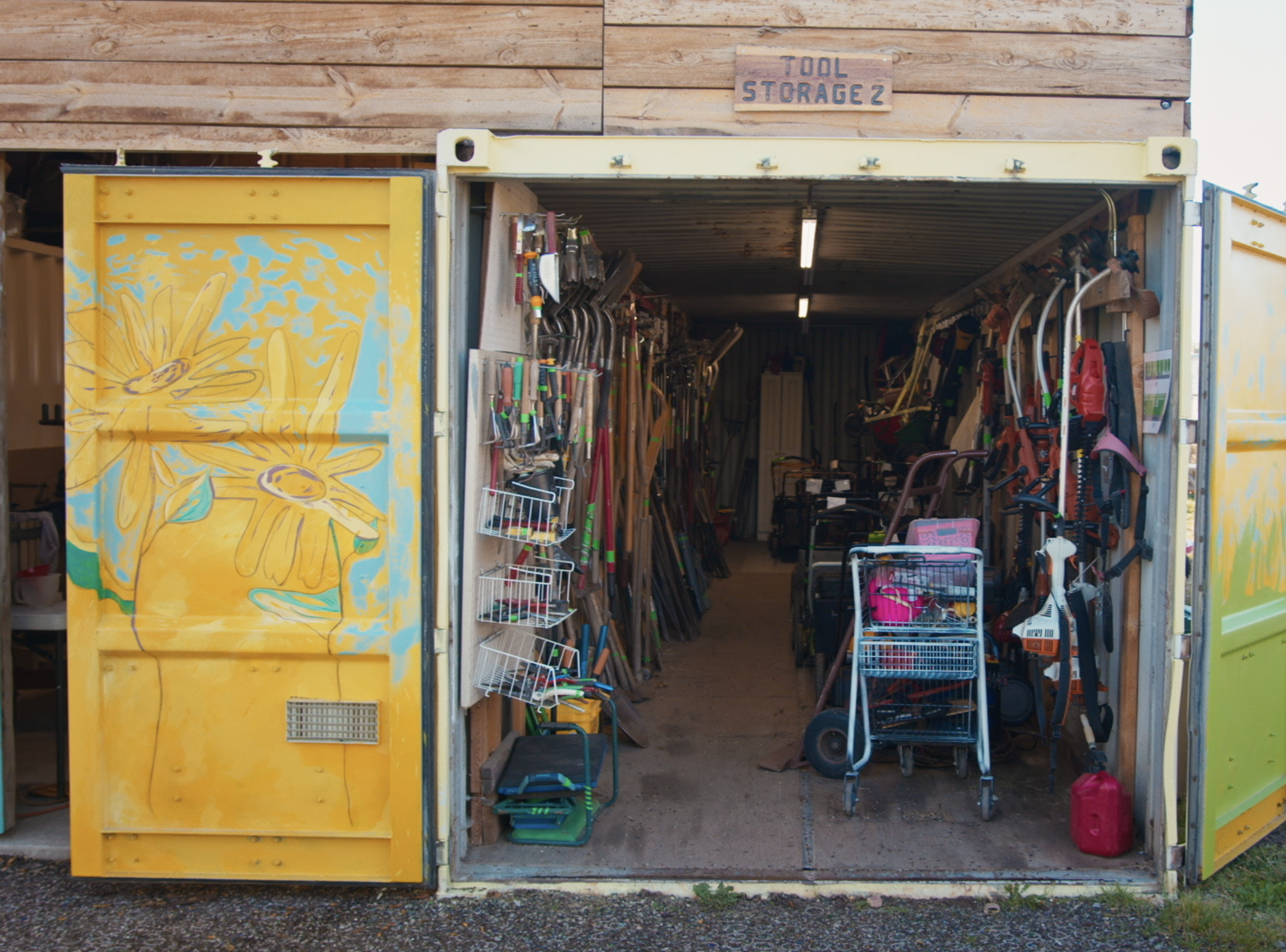 A storage shed with tools and gardening equipment organized on shelves and hanging on walls. The door is painted with yellow flowers. An overhanging sign reads "Tool Storage2.